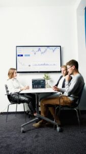 Servicios Three business professionals in a meeting room discussing financial charts displayed on a screen.